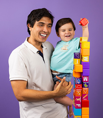 Marco smiling facing the camera with a young child stacking colorful toy blocks
